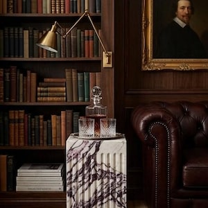 May include: A square white marble pedestal with deep purple veining, featuring a fluted design. On top sits a clear glass decanter filled with amber liquid and two matching crystal tumblers with a diamond-cut pattern on a silver tray.