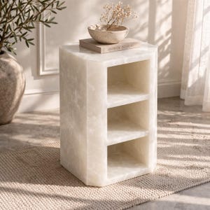 May include: A white, hexagonal side table with three open shelves. The table is made of a light-colored, veined material. A small bowl with dried flowers and a book with the text "Aesthetic Interiors" are on top.