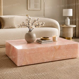 May include: A rectangular coffee table crafted from polished rose quartz, featuring a smooth, translucent pink surface with natural stone veining. The low-profile, block-style table sits directly on the floor, showcasing a solid, heavy construction.