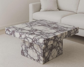 May include: Square marble coffee table with a white and purple veined pattern. The table has a solid square base and a matching square top. The table is in a modern style and is placed in a living room setting.