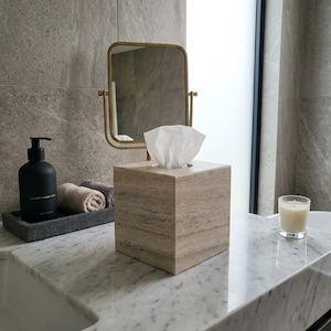 Puede incluir: Escena de baño con encimera de mármol. Cuenta con una caja de pañuelos cuadrada, un espejo con marco dorado y un dispensador de jabón negro. Toallas enrolladas y una vela encendida crean un ambiente relajante.