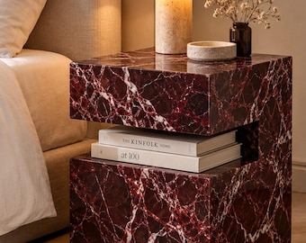 Puede incluir: Una mesa auxiliar cúbica hecha de mármol rojo intenso con vetas blancas prominentes. La mesa cuenta con un estante central abierto que contiene dos libros apilados titulados The Kinfolk Home y at 100, coronada por una lámpara.