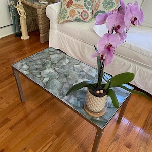 May include: A rectangular coffee table with a stone-like, gray-green top and a silver metal frame. A potted orchid with pink flowers and green leaves sits on the table. The table is 48 inches long and 24 inches wide.