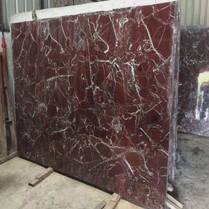 May include: Large, polished slabs of red marble with white veining. The marble is stacked, showcasing the natural patterns and textures. The slabs are supported by wooden beams.