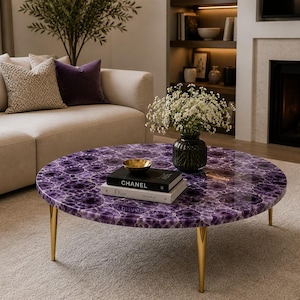 May include: A round coffee table with a polished purple surface, supported by three gold-colored legs. The table has a glossy finish and features a decorative amethyst pattern. A stack of books and a small gold bowl are on top.