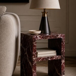 May include: A contemporary side table with a red marble effect and a lower shelf. A black lamp with a beige lampshade sits on top, alongside a small, light-coloured bowl. The table is next to a beige sofa.
