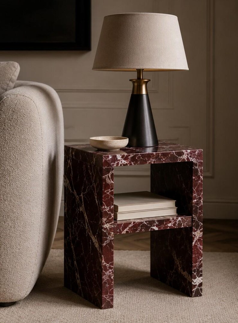 May include: A contemporary side table with a red marble effect and a lower shelf. A black lamp with a beige lampshade sits on top, alongside a small, light-coloured bowl. The table is next to a beige sofa.