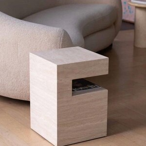 May include: A light beige, square side table with a magazine slot. The table is made of a light-colored stone material and has a minimalist design. The table is approximately 20 inches tall and 18 inches wide.