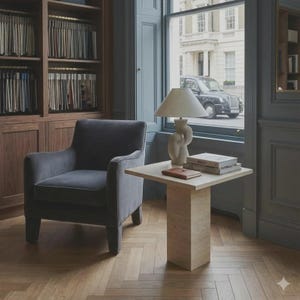 Op de afbeelding: Een gezellig interieur met een grijze fauteuil, een kleine vierkante bijzettafel en een decoratieve lamp. Op de tafel liggen boeken en een notitieboekje. Een houten boekenkast en een raam met uitzicht op de straat en een zwarte taxi maken de scène compleet.