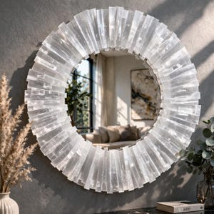 Peut inclure: Un miroir rond décoratif avec un cadre blanc texturé. Le cadre est composé de pièces rectangulaires disposées en motif radial. Le miroir reflète une pièce avec une fenêtre, un canapé et des œuvres d'art. Le miroir est fixé sur un mur gris.