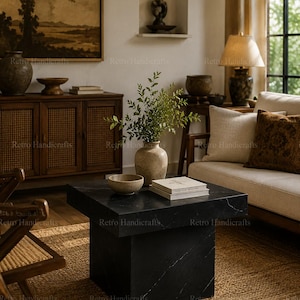 May include: A square black marble coffee table with white veining, featuring a thick tabletop and a solid block base. On the surface sits a beige ceramic vase with green foliage, a small matching bowl, and two stacked white books.