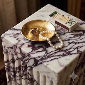 May include: A gold-toned circular dish containing a gold signet ring and a white pearl necklace, positioned next to a rectangular matchbox with a floral illustration, all resting on a white marble surface with deep purple veining.
