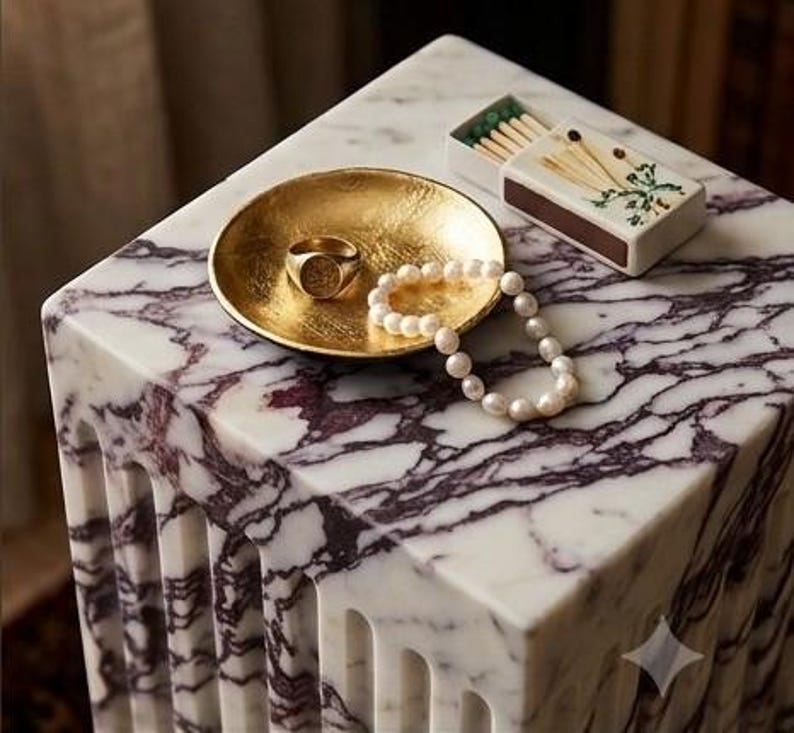 May include: A gold-toned circular dish containing a gold signet ring and a white pearl necklace, positioned next to a rectangular matchbox with a floral illustration, all resting on a white marble surface with deep purple veining.