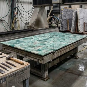 May include: A large, rectangular slab of green aventurine mosaic stone, likely for countertops or other surfaces, is being worked on by a large industrial machine. The stone is composed of many irregular pieces. A sign reads "Premium Aventurine Mosaic - Factory Grade."