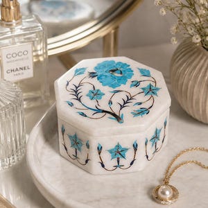 May include: Octagonal white marble jewellery box with a blue floral inlay. The box has a detailed pattern of flowers and leaves. A gold necklace with a pearl pendant is also visible.