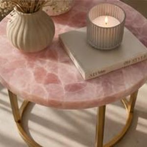 K&ouml;nnte beinhalten: Ein runder Beistelltisch mit einer Tischplatte aus rosa Quarz und goldfarbenen Beinen. Eine wei&szlig;e Vase mit Trockenblumen, ein Buch und eine brennende Kerze in einem Glashalter stehen auf dem Tisch. Die Tischoberfl&auml;che hat ein mosaikartiges Aussehen.