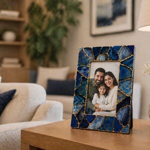 Op de afbeelding: Een rechthoekige fotolijst met een blauw en goud mozaïekontwerp. De lijst bevat een familiefoto. De lijst staat op een houten tafel in een woonkamer.