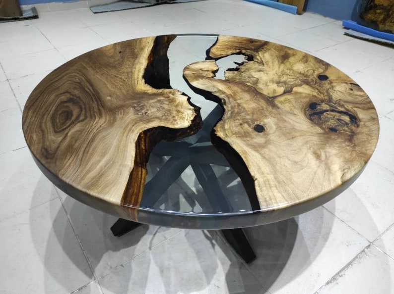 May include: A circular coffee table with a distinctive design. The tabletop is crafted from light brown wood, featuring dark brown and clear resin details. The table is supported by a dark brown base with four legs.