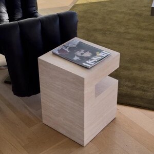 May include: A light-colored, square side table with a cutout on one side. A magazine with the word "ISSUE" is on top. The table is in a room with a black chair and a green rug.