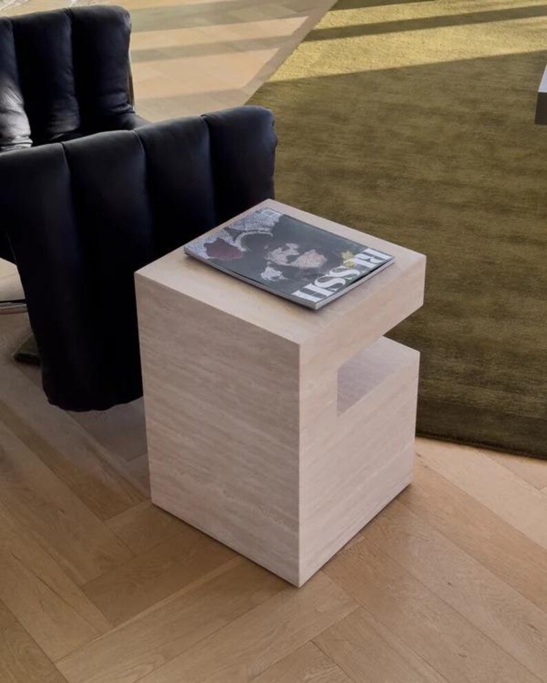 May include: A light-colored, square side table with a cutout on one side. A magazine with the word "ISSUE" is on top. The table is in a room with a black chair and a green rug.