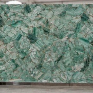 May include: Large rectangular slab of green and white stone, composed of irregular, interlocking pieces. The stone has a polished surface and is supported by wooden beams. The overall appearance is of a mosaic or a natural gemstone.