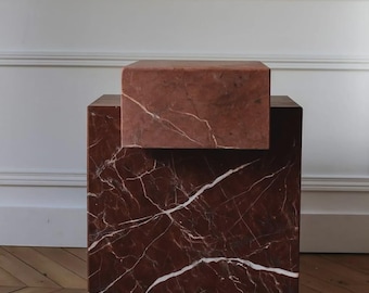 Puede incluir: Una mesa auxiliar de dos niveles de mármol rojo con vetas blancas. La sección inferior es un cubo grande, y la sección superior es un bloque rectangular más pequeño. La mesa está sobre un suelo de madera.