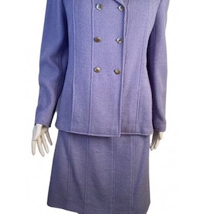 Puede incluir: Un traje de chaqueta y falda de color lavanda de doble botonadura. La chaqueta tiene un cuello de muesca y la falda es de largo hasta la rodilla.