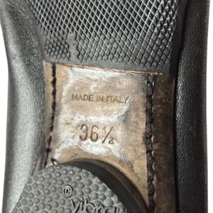 May include: The underside of a black leather shoe featuring a textured black rubber Vibram sole. The center of the sole is light brown with the text MADE IN ITALY and the size 36.5 clearly visible. A circular Vibram logo is embossed on the heel.