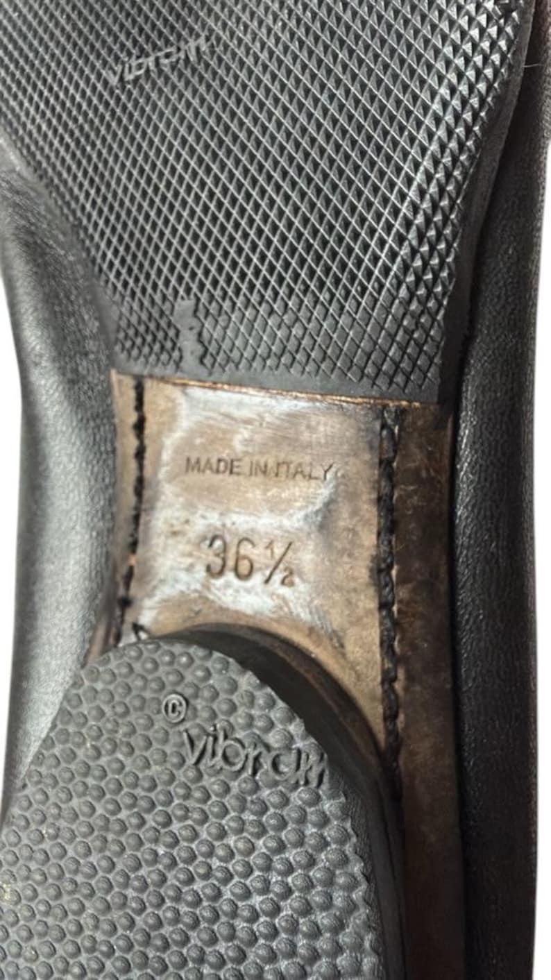 May include: The underside of a black leather shoe featuring a textured black rubber Vibram sole. The center of the sole is light brown with the text MADE IN ITALY and the size 36.5 clearly visible. A circular Vibram logo is embossed on the heel.