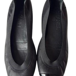 May include: A pair of black leather ballet flats featuring a rounded toe with a glossy black patent leather cap. The shoes have a black elasticized trim around the opening, a small pull tab at the heel, and the Chanel logo printed on the inner sole.