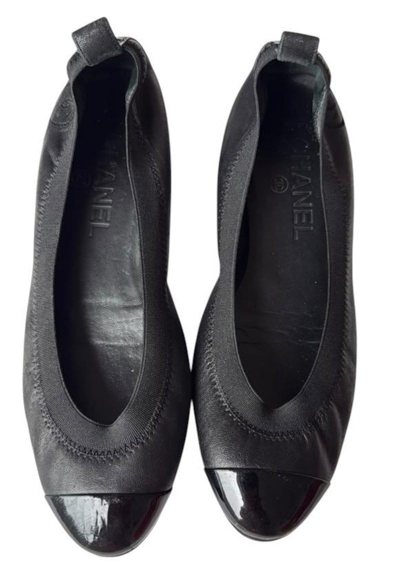 May include: A pair of black leather ballet flats featuring a rounded toe with a glossy black patent leather cap. The shoes have a black elasticized trim around the opening, a small pull tab at the heel, and the Chanel logo printed on the inner sole.