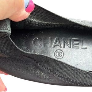 May include: A black leather ballet flat with a black elasticized trim. The interior insole is black and features the embossed text CHANEL, the interlocking CC logo, and the words MADE IN ITALY stamped in a circular pattern on the heel area.