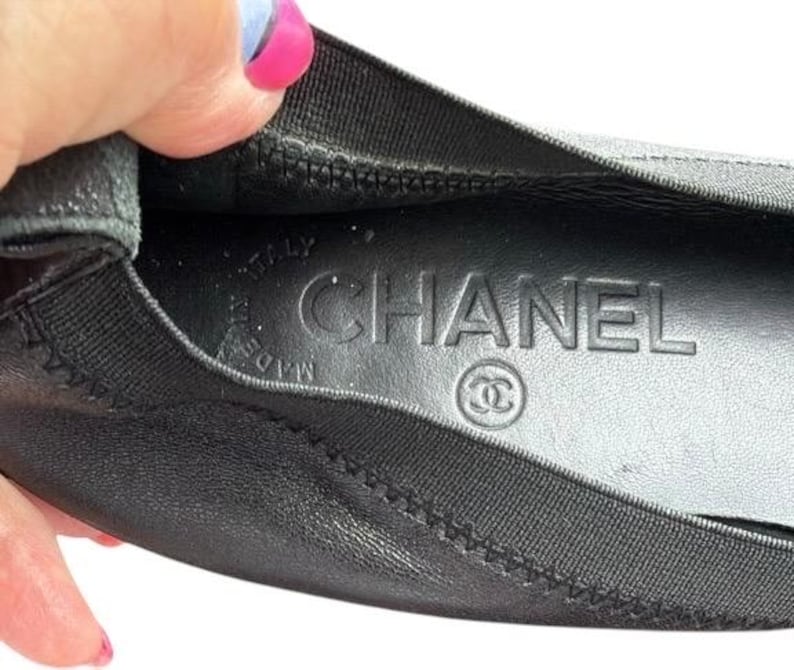 May include: A black leather ballet flat with a black elasticized trim. The interior insole is black and features the embossed text CHANEL, the interlocking CC logo, and the words MADE IN ITALY stamped in a circular pattern on the heel area.