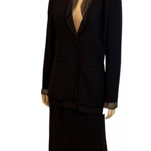 Puede incluir: Un traje de chaqueta y falda negro con un cuello de gasa negro. La chaqueta tiene un cierre de botón único y dos bolsillos. La falda es de largo hasta la rodilla.