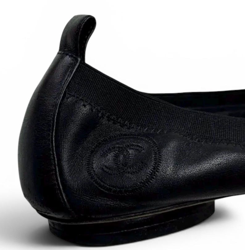 May include: A black leather ballet flat featuring a rounded heel tab and a black elasticized topline. The side of the shoe displays a circular embossed logo with the interlocking CC emblem. The construction includes visible stitching along the heel.