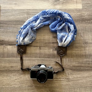 May include: A silver camera with a blue and white floral patterned camera strap. The camera has a black lens and the text "minolta" printed on the top.