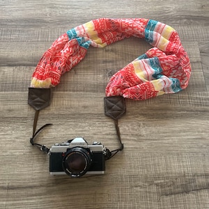May include: A vintage silver camera with a black lens and the word "minolta" printed on it. The camera is attached to a colorful patterned fabric camera strap with brown leather accents.