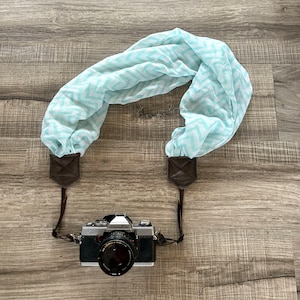 May include: A light blue and white patterned camera strap made of fabric with brown leather accents. The strap is attached to a silver camera with a black lens. The camera has the word "minolta" printed on it.