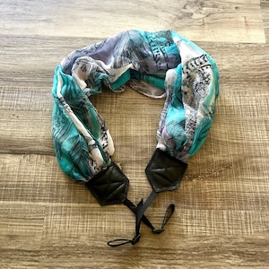 Puede incluir: Una correa de cámara estampada con un diseño turquesa, blanco y morado. La correa tiene extremos de cuero negro y cordones negros ajustables. La correa está hecha de una tela ligera.