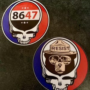 May include: Two round stickers. One features a skull with the number "8647" inside a red, white, and blue circle. The other sticker shows a bear wearing a hat that says "RESIST" inside a similar circle.