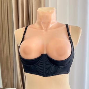 Könnte beinhalten: Schwarzes Dessous-Set auf einer Schaufensterpuppe. Das Set besteht aus einem Demi-Cup-BH mit verstellbaren Trägern und passender Hüfthose. Der BH hat einen gewellten Rand und die Unterwäsche ein dekoratives Schnürdetail vorne.