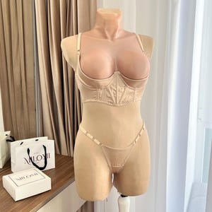 May include: A beige lingerie set consisting of a structured, open-cup corset top with vertical stitching and adjustable straps, paired with matching high-cut thong underwear featuring thin side straps with small gold-toned metal ring hardware details.