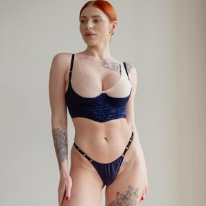 Op de afbeelding: Een marineblauwe lingerie set. De satijnen bustier heeft beige cups en zwarte bandjes. De bijpassende string heeft goudkleurige accenten. De bustier heeft een gestructureerd ontwerp.