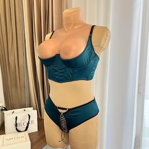 May include: Teal lingerie set on a mannequin. The set includes a satin bra with black straps and a matching pair of shorts with a lace-up detail. The bra has underwire support. The shorts have black trim.