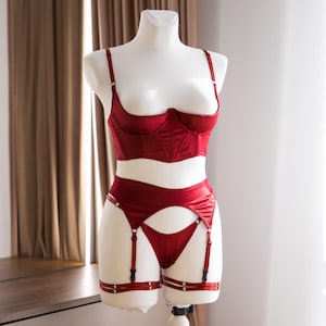 Op de afbeelding: Een rode satijnen lingerie set met een bustier, jarretelgordel en kousen. De set heeft delicate kantdetails en verstelbare bandjes.