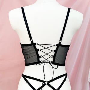 Op de afbeelding: Zwarte kanten lingerie top met een veter aan de achterkant en verstelbare bandjes. De top is gemaakt van een transparante mesh stof en heeft een delicaat bloemenpatroon.