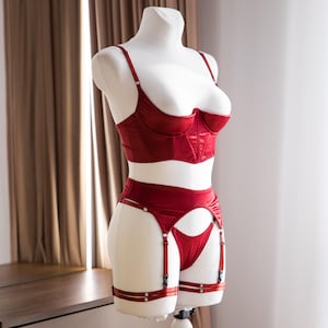 Op de afbeelding: Een rode lingerie set op een mannequin. De set bestaat uit een bh met verstelbare bandjes, een hoge taille jarretelgordel en een bijpassende string. De lingerie is gemaakt van glanzend materiaal en heeft een gestructureerd ontwerp.