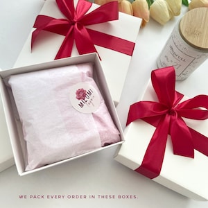 K&ouml;nnte beinhalten: Zwei wei&szlig;e Geschenkboxen mit roten Schleifen. Eine Box hat ein rosa Seidenpapierfutter mit einem runden Aufkleber, auf dem "MIELOMI by me" steht. Die andere Box enth&auml;lt eine Kerze mit einem Holzdeckel und einem Etikett mit der Aufschrift "MINDFULL CANDLE NO. 21".