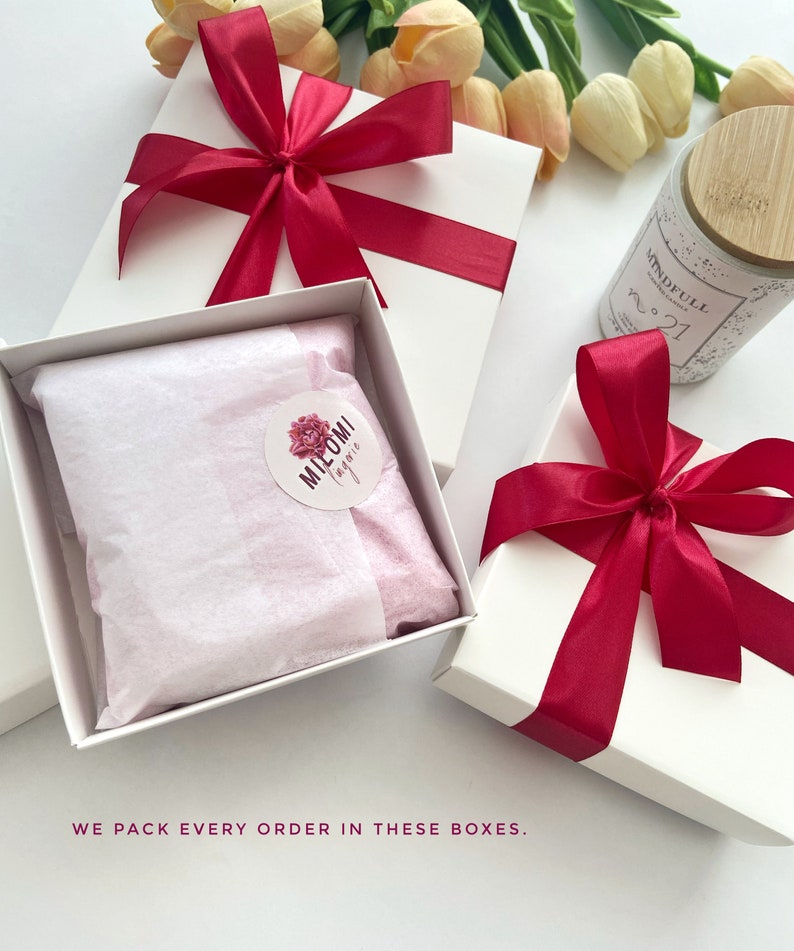 K&ouml;nnte beinhalten: Zwei wei&szlig;e Geschenkboxen mit roten Schleifen. Eine Box hat ein rosa Seidenpapierfutter mit einem runden Aufkleber, auf dem "MIELOMI by me" steht. Die andere Box enth&auml;lt eine Kerze mit einem Holzdeckel und einem Etikett mit der Aufschrift "MINDFULL CANDLE NO. 21".