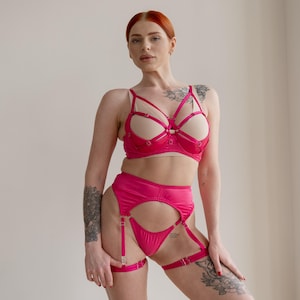 Op de afbeelding: Een felroze lingerieset met een beha met beige cups en een hoog getailleerde jarretelgordel met jarretels. De beha heeft een uniek bandjesontwerp. De set is gemaakt van een gladde, glanzende stof.